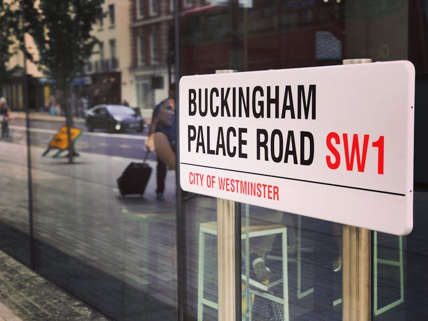 A street scene showing a white public signboard with black and red text indicating 'Buckingam Place Road SW1' in Westminster, London. The sign is mounted on metal poles positioned on the pavement outs